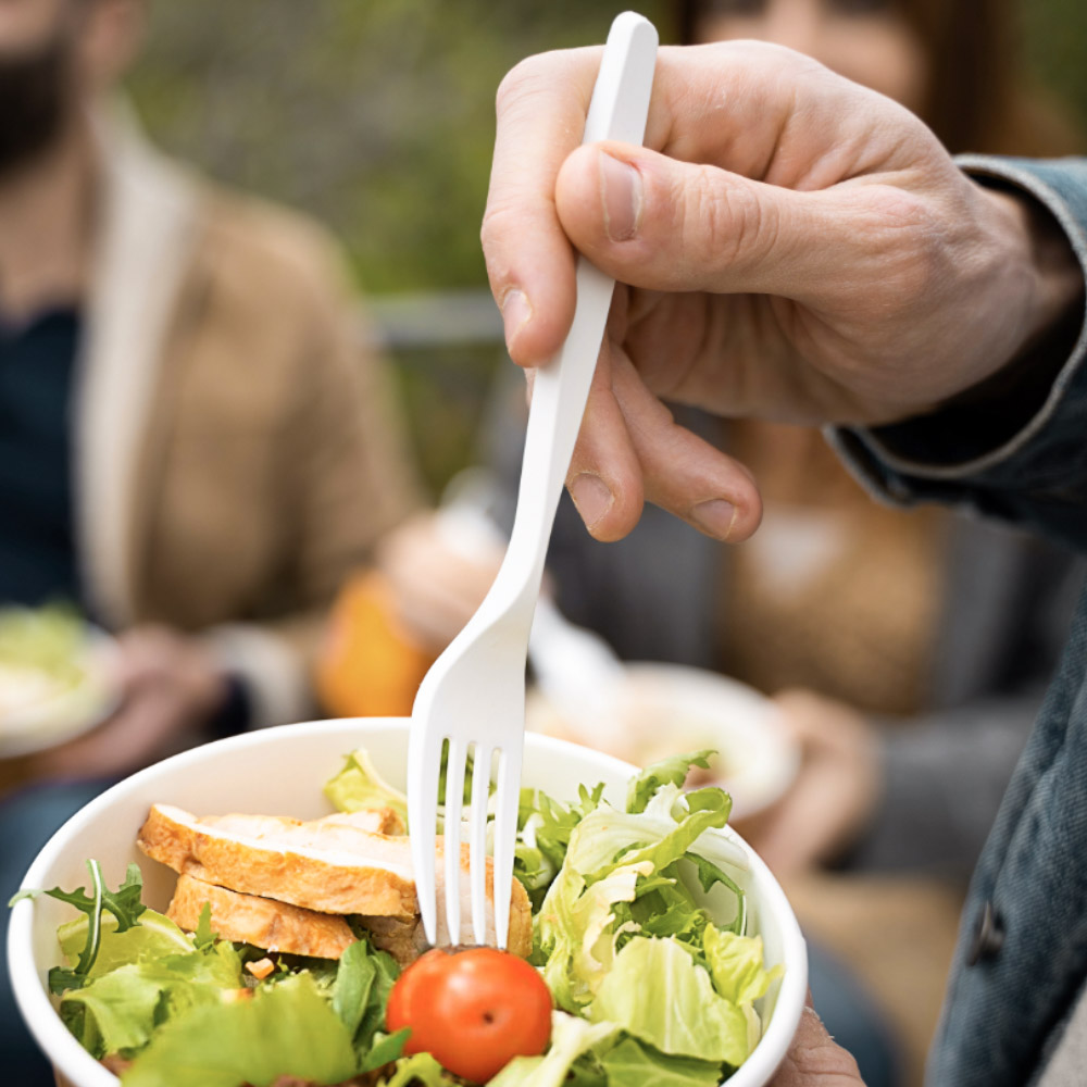 Forchetta compostabile in bioplastica utilizzata per gustare un’insalata con pollo e pomodoro, in un contesto conviviale e sostenibile all’aperto.