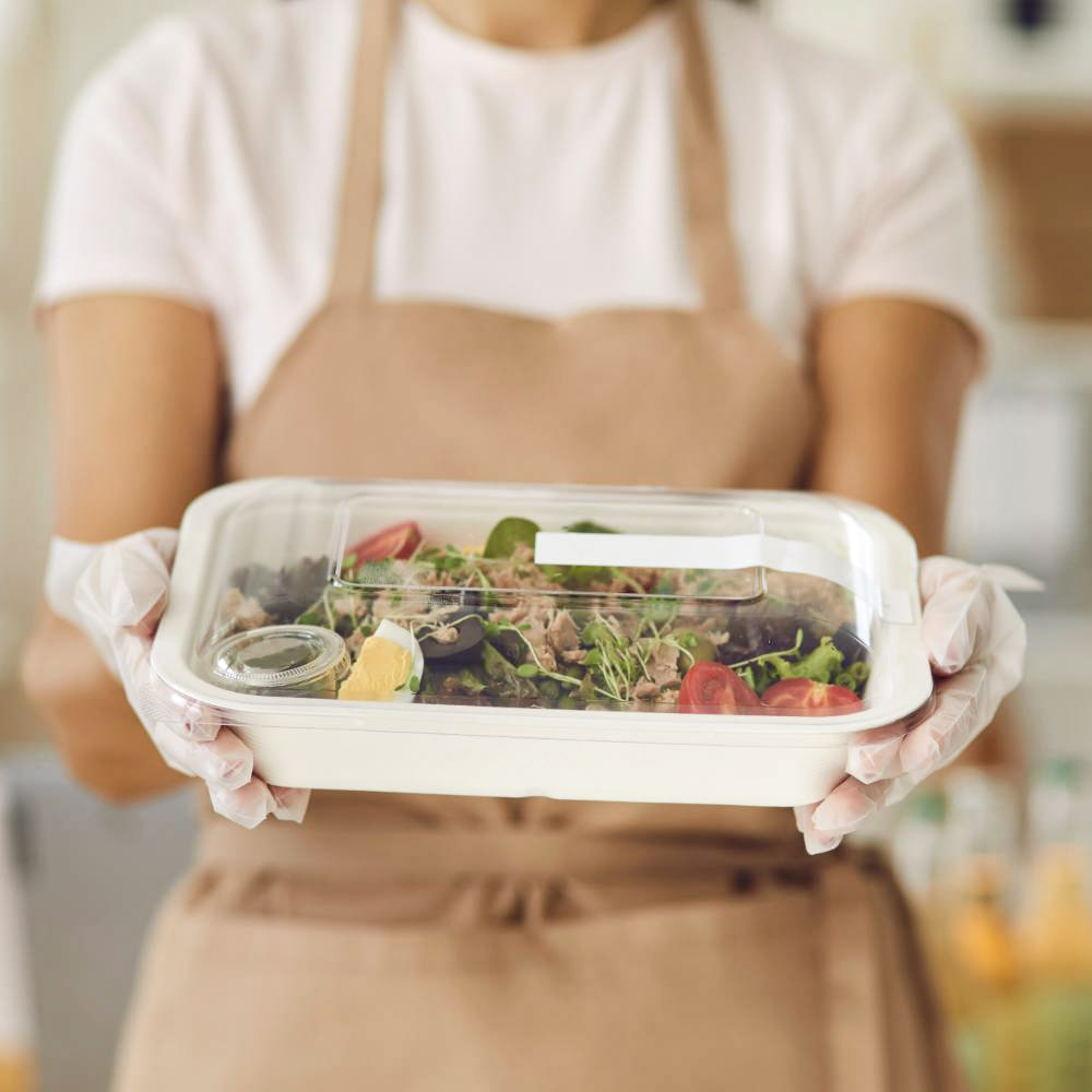 Operatore mensa con guanti che porge una vaschetta compostabile con coperchio trasparente, contenente un'insalata fresca con ingredienti naturali.